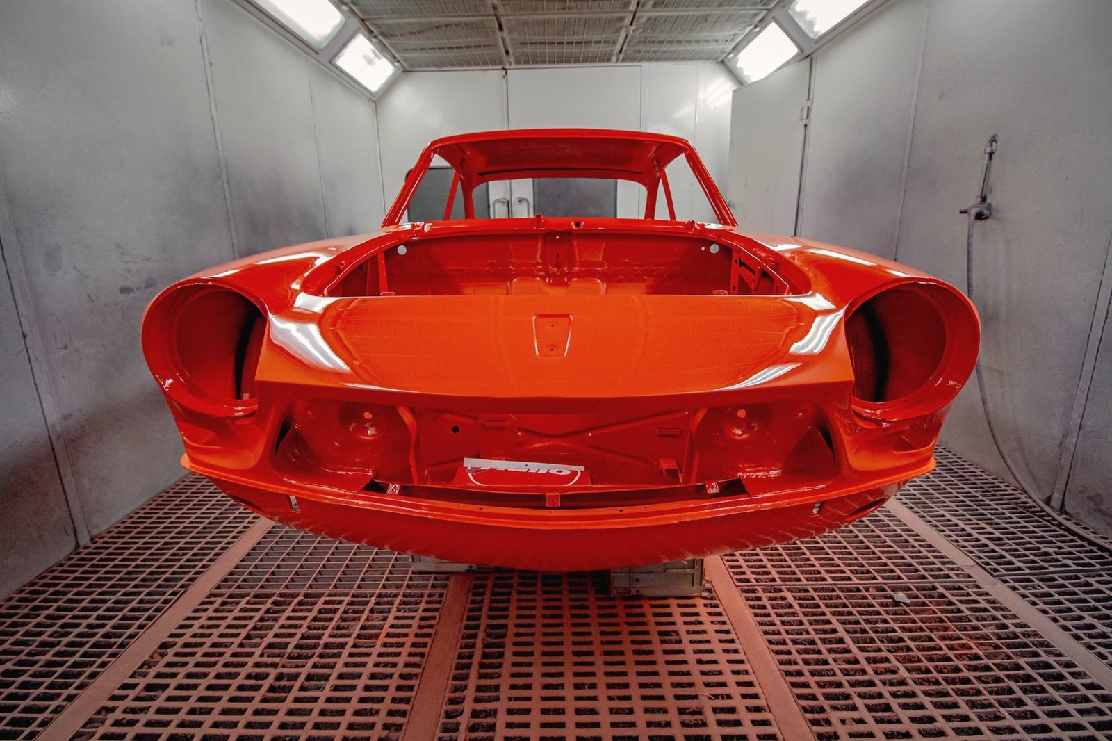 A bare car body shell freshly painted bright red-orange, standing on the grated floor of a paint booth