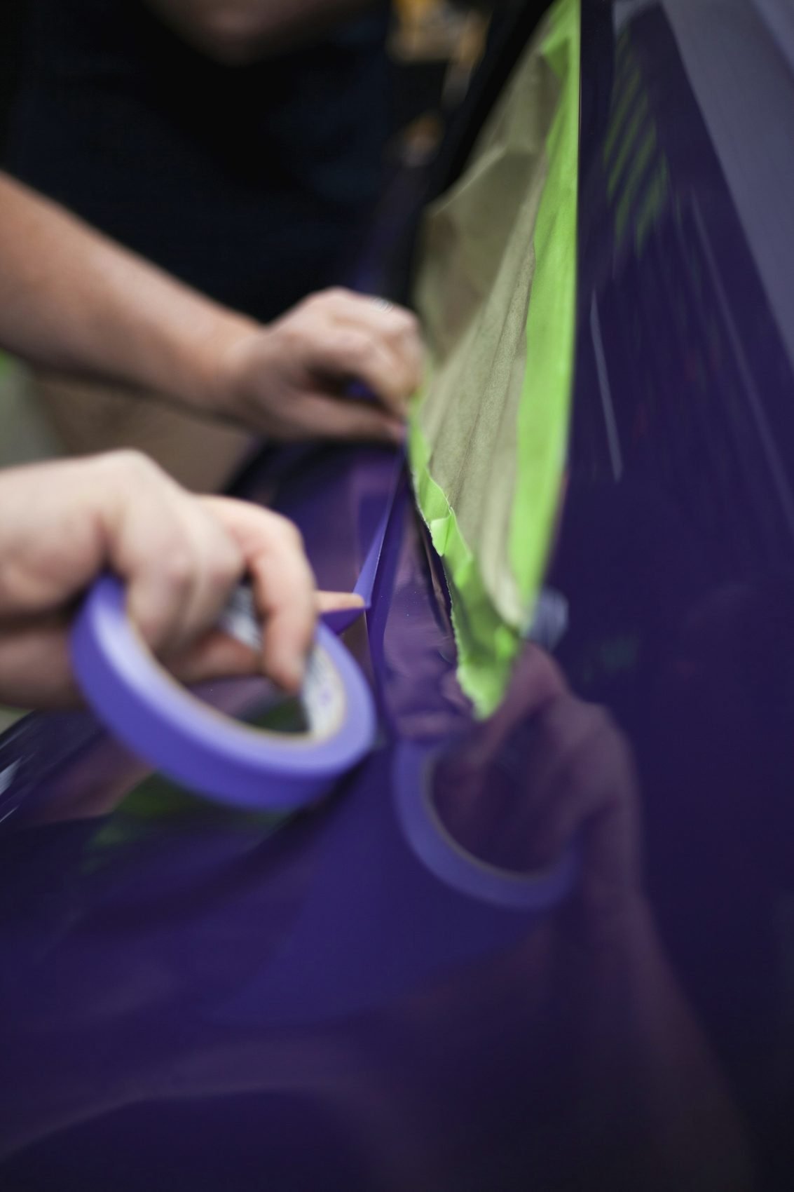 Hands laying blue masking tape along the edge of a purple car panel