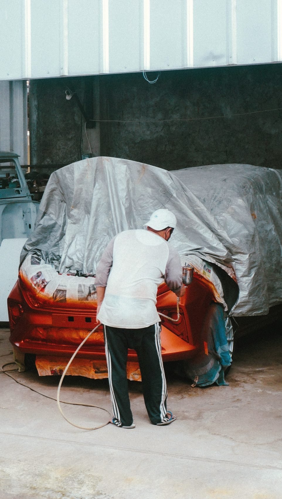 A person with their back to the camera sanding the front of an orange car under a dust sheet
