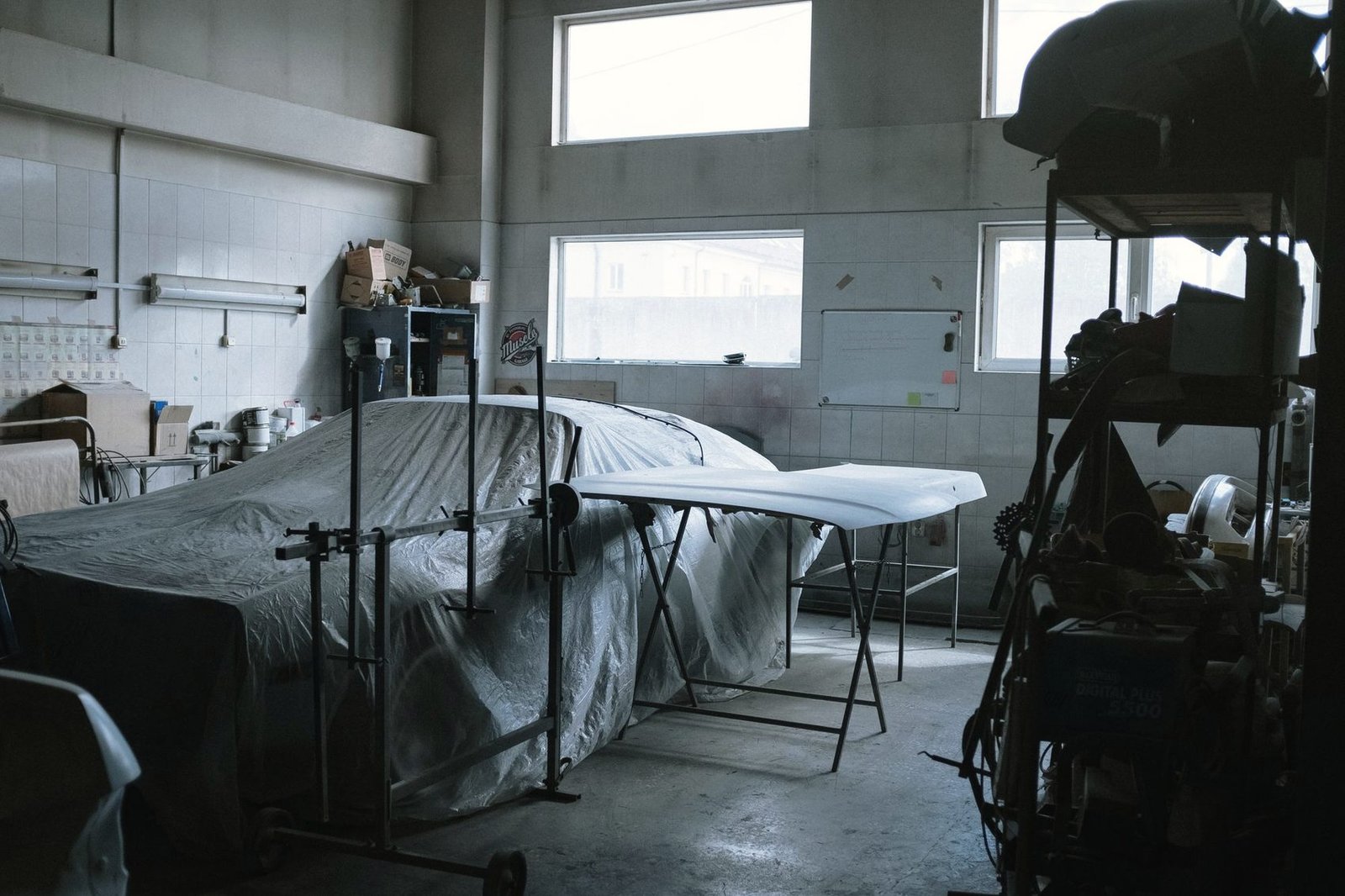 A car under a dust sheet in a workshop, with a loose panel resting on a stand beside it