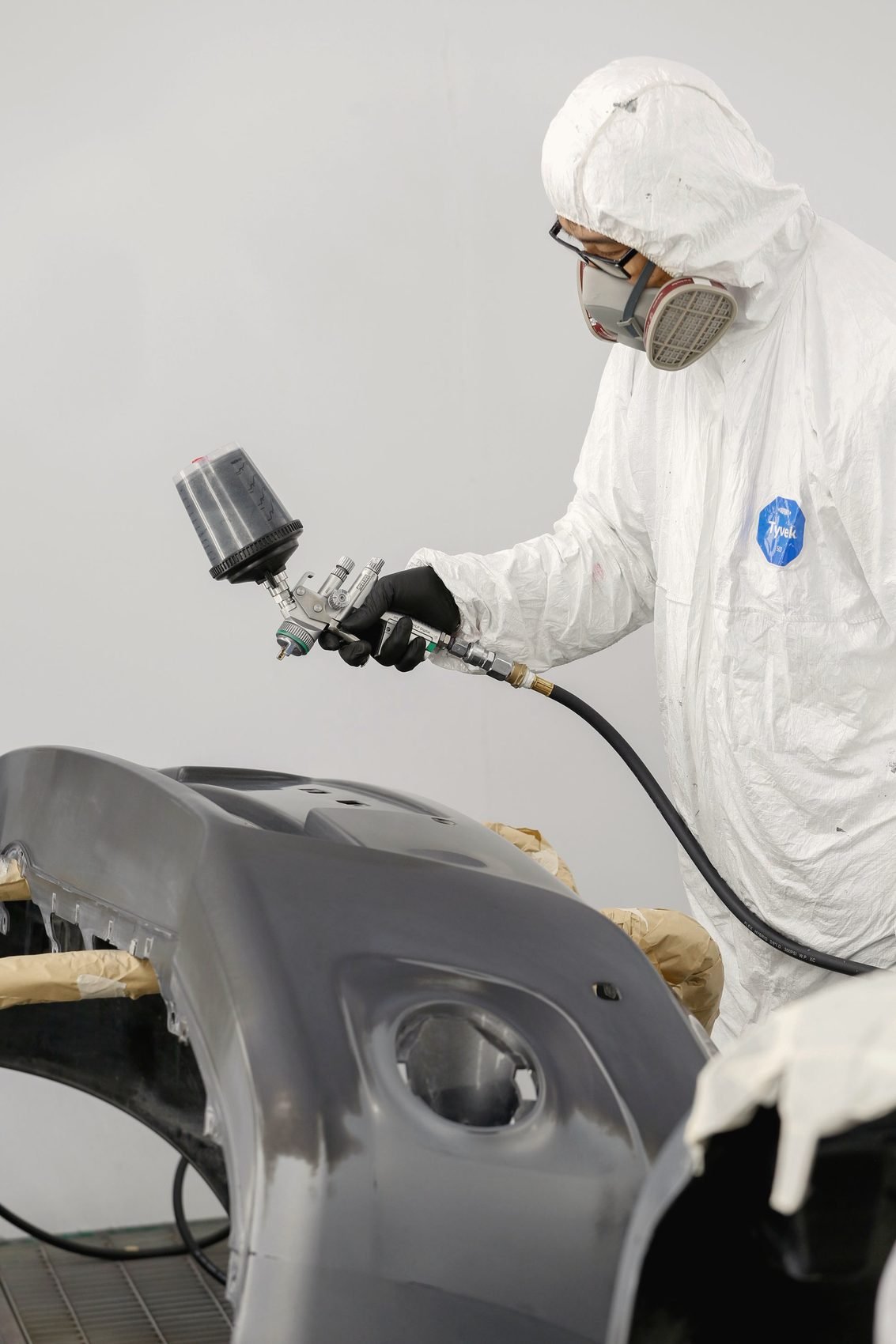 A painter in a white suit and respirator spraying a grey bumper cover
